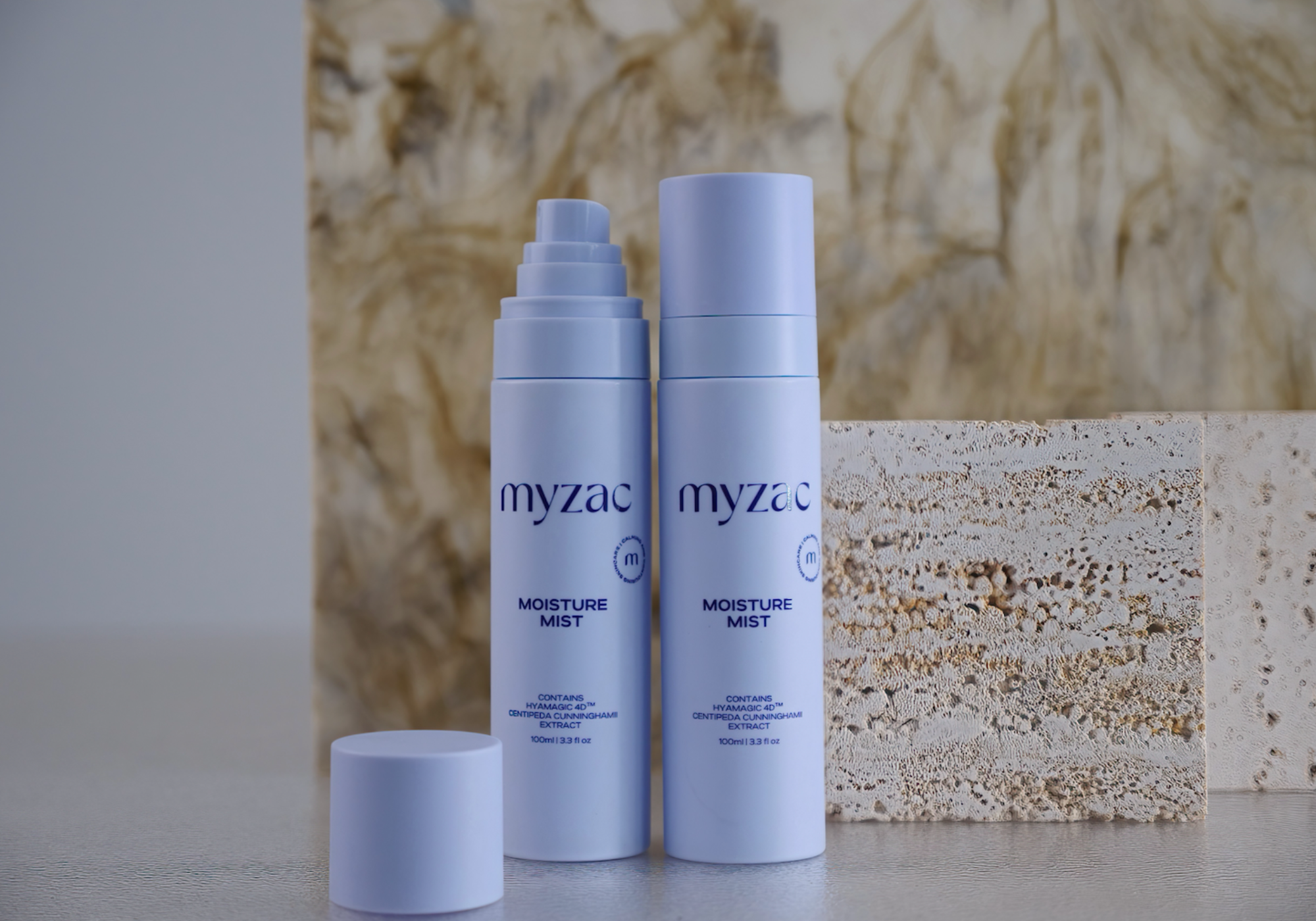Two MYZAC Moisture Mist bottles with one opened to reveal the pump, styled with textured stone backdrops, highlighting the product’s clean and clinical design.